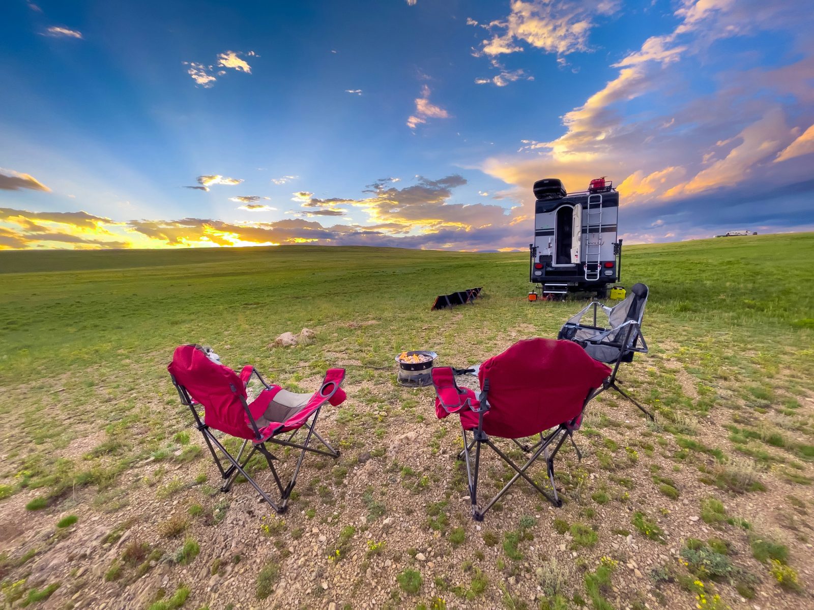 Two red camping chairs around a fire pit on open grassland at sunset, with a truck camper parked behind, black and white.