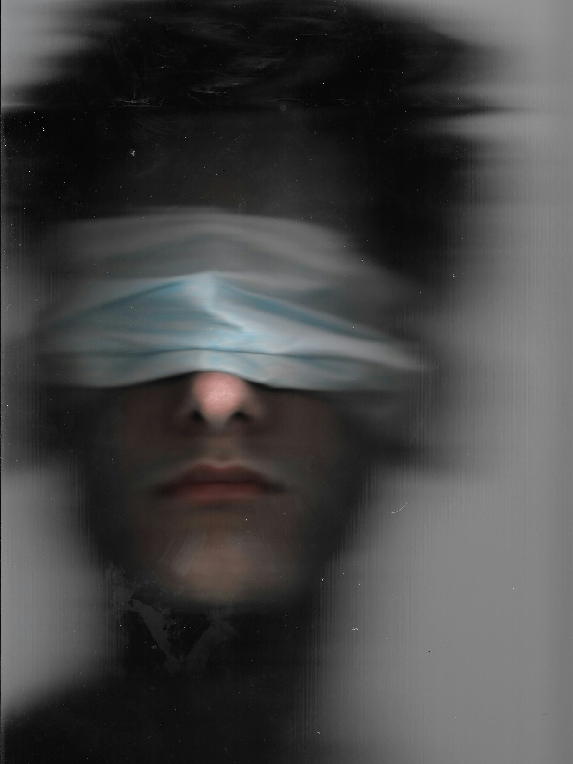 A blurred portrait of a man with a cloth tied over his eyes, black and white.